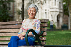 Virginia M.-Y. Lee, PhD, sits on a campus wooden bench, smiling and relaxed legs crossed, wearing blue pants and a multicolored shirt.